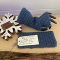 A pair of denim blue fingerless wrist warmers displayed on a wooden surface, with a snowflake decoration in the background.