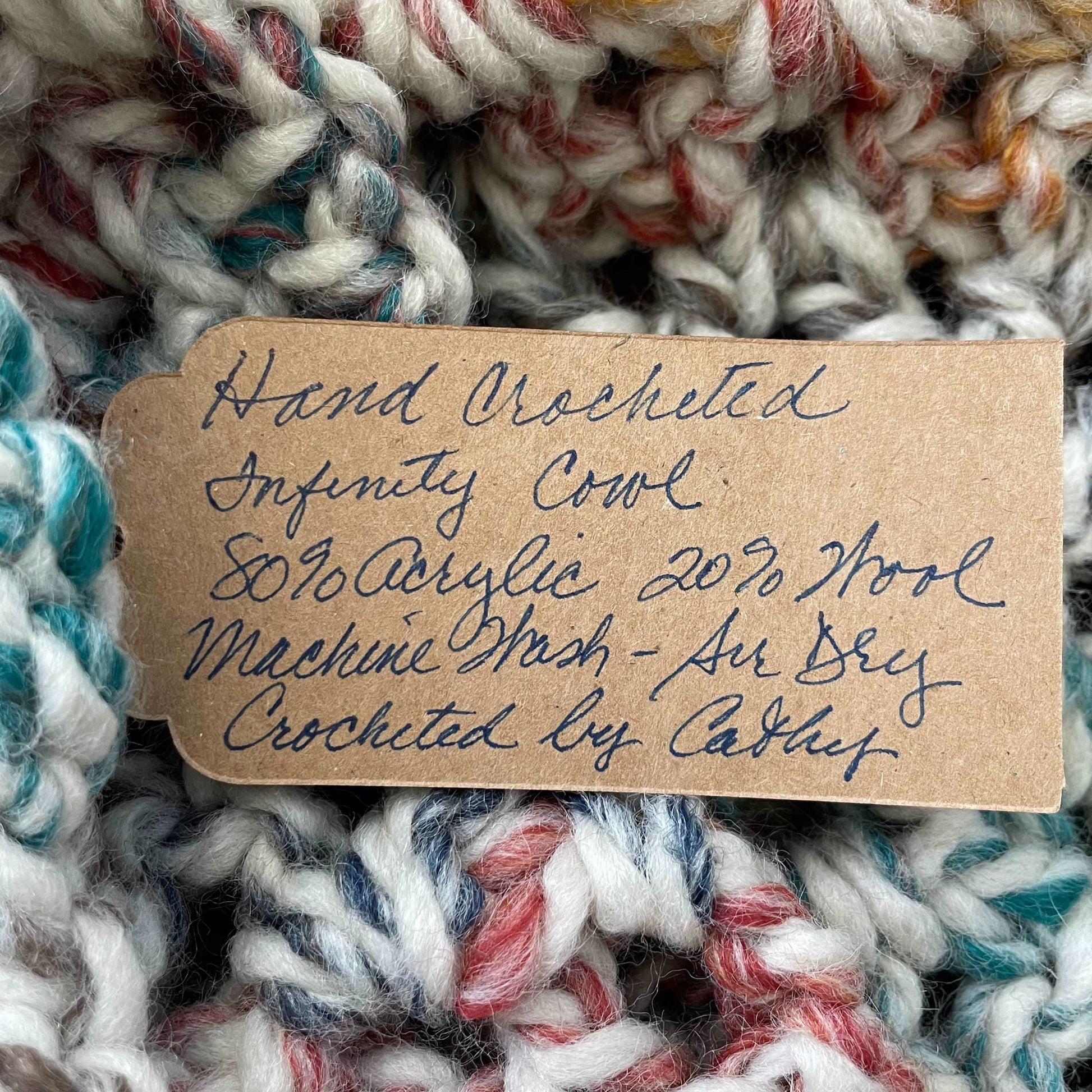 close up of handwritten tag reading hand crocheted infinity cowl 80% acrylic 20% wool machine wash air dry crocheted by cathy